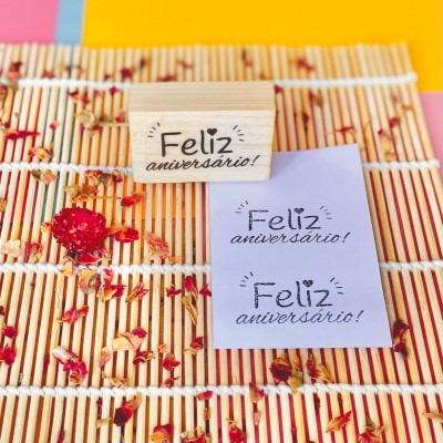 Carimbo de madeira com texto 'Feliz aniversário!' em esteira de bambu com papel e pétalas secas