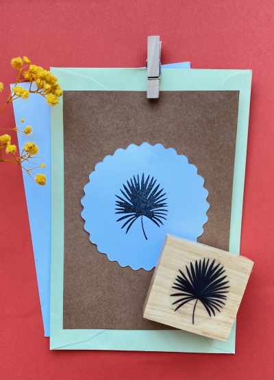 Cartão castanho com selo azul e folha preta, carimbo de madeira, envelope verde e flores amarelas