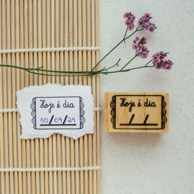 Carimbo de madeira com texto Hoje é dia, papel com mesmo texto e data, flores roxas e tapete de bambu