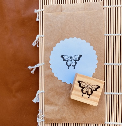 Carimbo de madeira com imagem de borboleta e embalagem de papel castanho com etiqueta branca, sobre base de palha