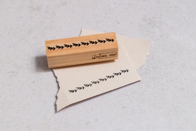 Carimbo de madeira com desenho de ramos e texto 'untone one' sobre papel rasgado