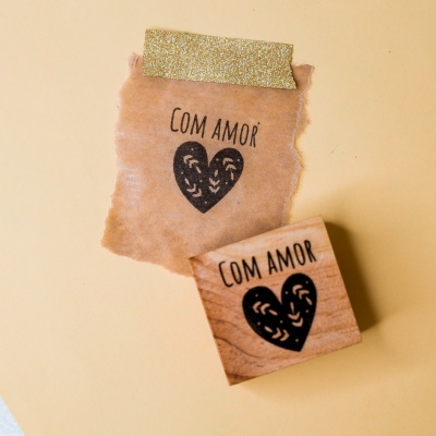 Carimbo de madeira com texto 'COM AMOR' e coração preto numa folha de papel castanho colada com fita dourada