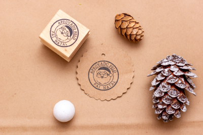 Selo e carimbo 'APROVADO PELO PAI NATAL' com pinhas e esfera branca sobre superfície castanha