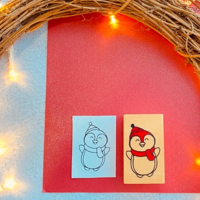 Dois carimbos de madeira com pinguins sorridentes sobre superfície vermelha com luzes e ramos secos