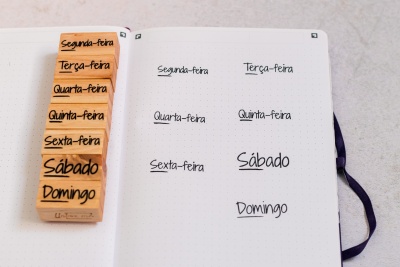 Caderno aberto com páginas pontilhadas e carimbos de madeira com nomes dos dias da semana em português.