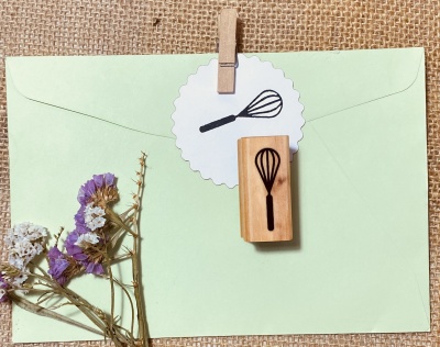 Carimbo de madeira com desenho de batedor de arames preso a etiqueta num envelope verde claro com flores secas ao lado