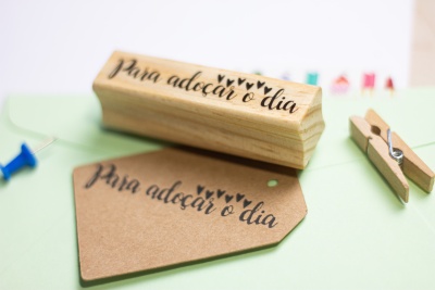 Carimbo de madeira com frase 'para adoçar o dia', papel kraft carimbado, alfinete azul e pregador de madeira