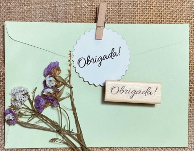 Envelope verde claro com cartão 'Obrigada!' e flores secas sobre tecido bege