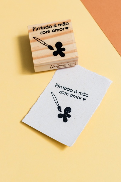 Carimbo de madeira com desenho e texto em preto sobre superfície amarela.