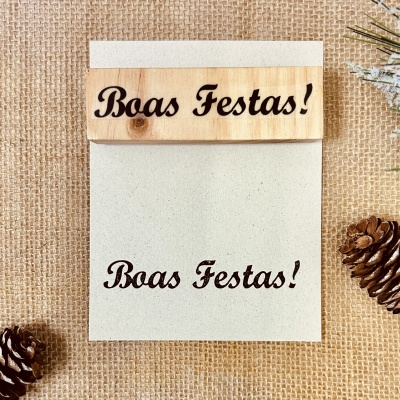 Cartão 'Boas Festas!' em madeira e papel sobre tecido juta com pinhas.