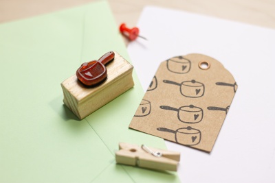 Carimbo de madeira com desenho de panela, etiqueta com padrão de panelas, pregador de madeira e envelope verde