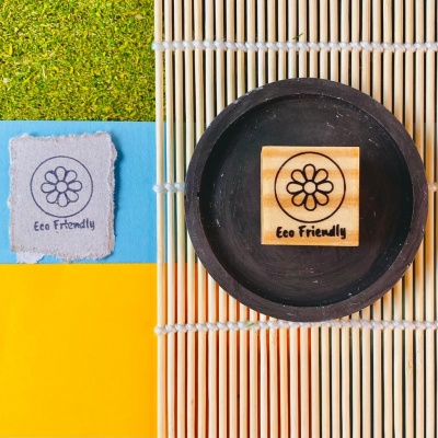 Carimbo de madeira com símbolo de flor e texto Eco Friendly em prato preto sobre base de bambu, papel colorido e grama artificial ao lado.