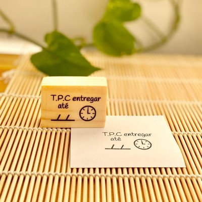 Carimbo de madeira com mensagem 'T.P.C entregar até' ao lado de folha carimbada sobre superfície de ripas de madeira