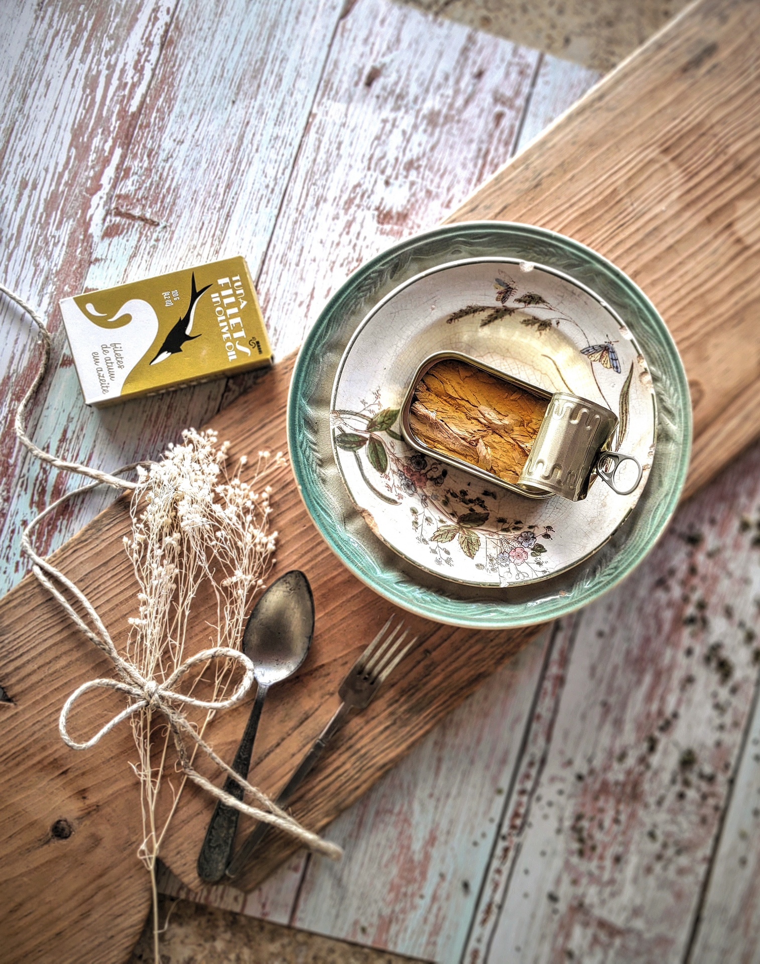 Prato com sardinhas, colher, garfo, caixa dourada, e ramo seco sobre tábua de madeira.