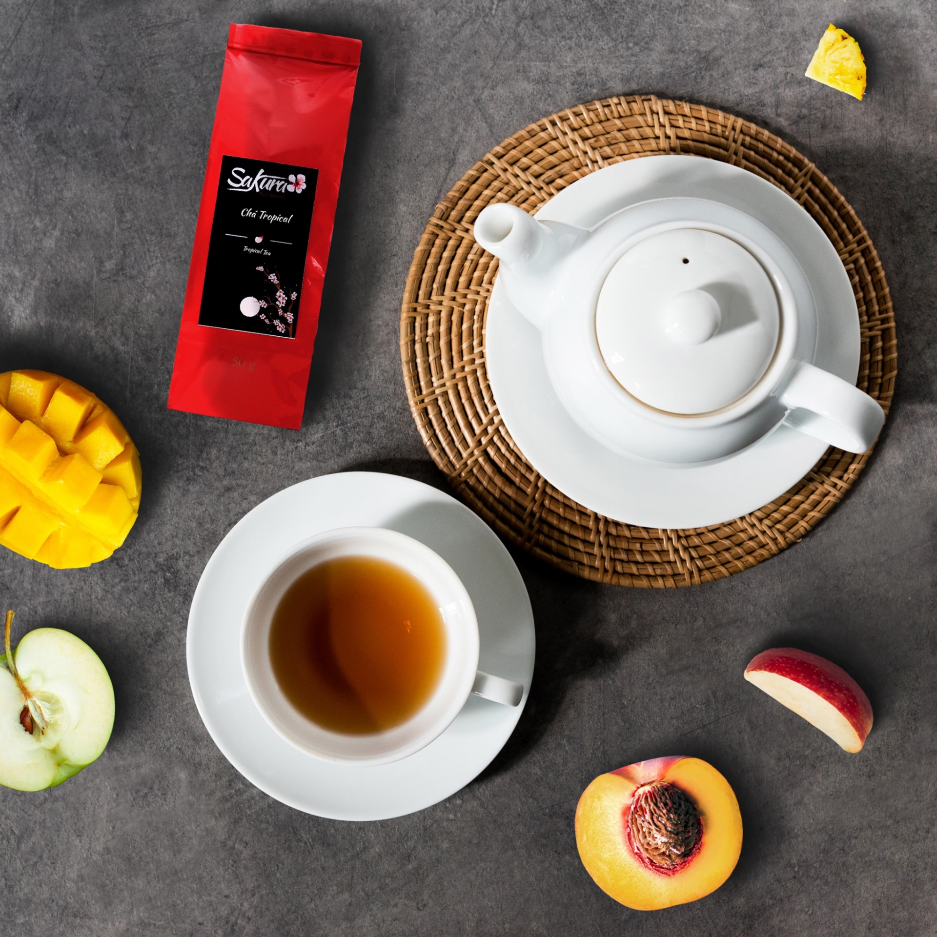 Infusão Tropical Pacote vermelho de chá, bule e chávena branca, frutas frescas em superfície cinzenta