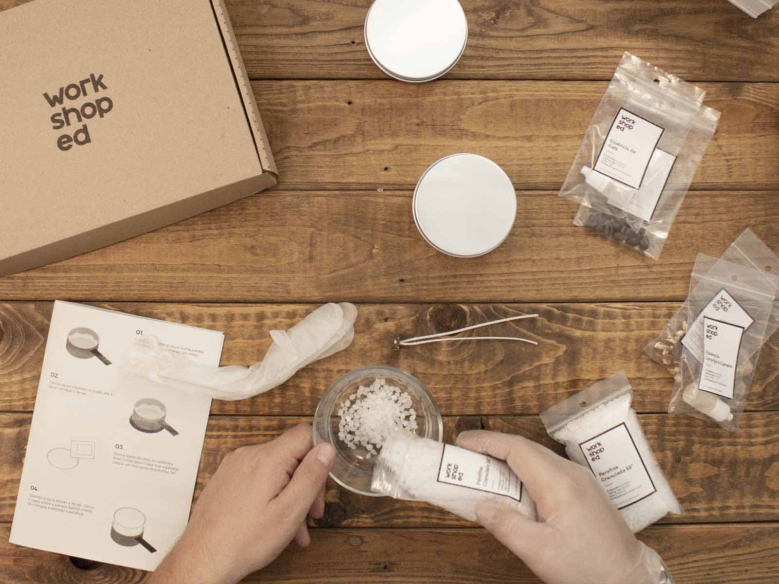 Workshop Box: Velas Aromáticas Kit DIY com caixa de cartão, recipientes brancos, sacos com etiquetas e manual sobre mesa de madeira.