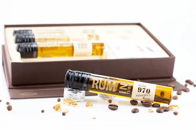 À Descoberta do Rum Português Frasco de rum em vidro com tampa preta e rótulo branco, em superfície branca com grãos de café e canela, ao lado de caixa aberta com outros frascos.