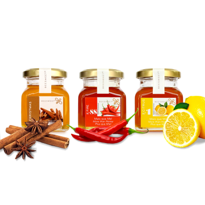 Três frascos de mel com tampas douradas, decorados com canela, malaguetas e limão, etiquetas diferentes em fundo branco.