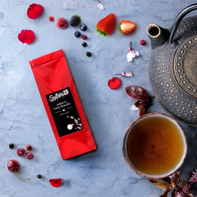 Pacote vermelho de chá Sakuma, chávena de chá castanho e bule preto com frutas vermelhas e pétalas numa superfície cinzenta