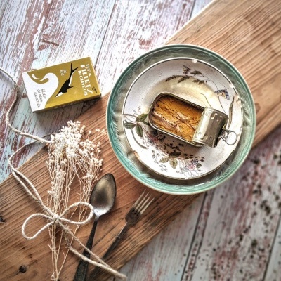 Prato com sardinhas, colher, garfo, caixa dourada, e ramo seco sobre tábua de madeira.