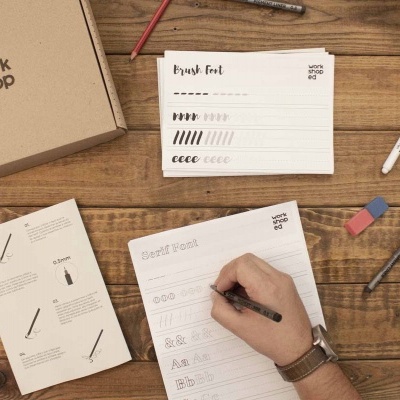 Mesa de madeira com caixa de cartão, folhas com tipos de letra e mão escrevendo com caneta.