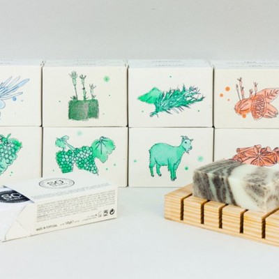 Sabonetes artesanais em embalagens brancas com ilustrações coloridas e uma barra de sabonete em base de madeira