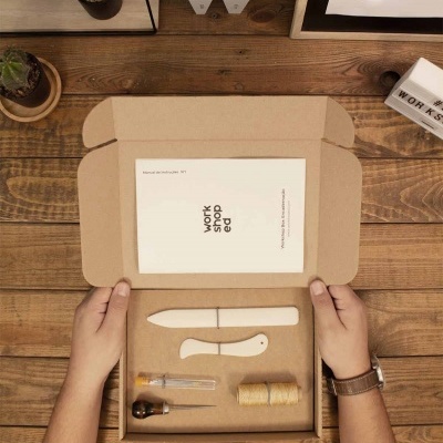 Kit de trabalho manual com faca branca, peça ergonômica, vidro com líquido, rolo de fio e livreto numa caixa de papelão sobre mesa de madeira.