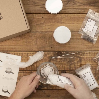 Kit DIY com caixa de cartão, recipientes brancos, sacos com etiquetas e manual sobre mesa de madeira.
