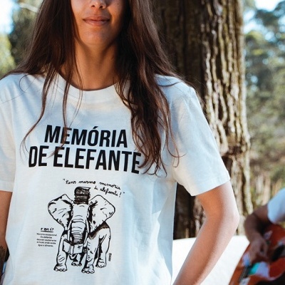 Pessoa com t-shirt branca estampada com elefante e texto MEMÓRIA DE ELEFANTE em ambiente de floresta