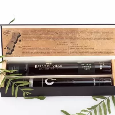 Conjunto embalagem com garrafa preta de vinho do Porto e caneta, dentro de caixa de madeira, com texto em português na tampa da caixa e folhas verdes em volta.