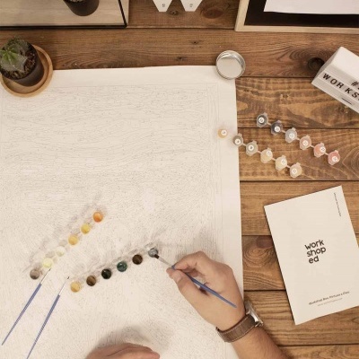 Pessoa a pintar quadro por números numa mesa de madeira com tintas, pincéis, folheto e plantas pequenas