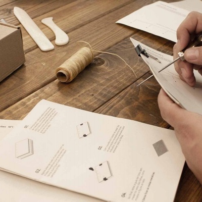 Mãos a costurar sapato branco com linha bege na mesa de madeira, com carretel de linha e instruções