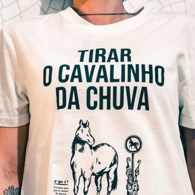 T-shirt branca com texto e ilustrações a preto, pessoa a usar, fundo de azulejos brancos e pretos