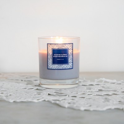 Vela aromática cinza em copo de vidro com etiqueta azul e branca sobre toalha rendada branca