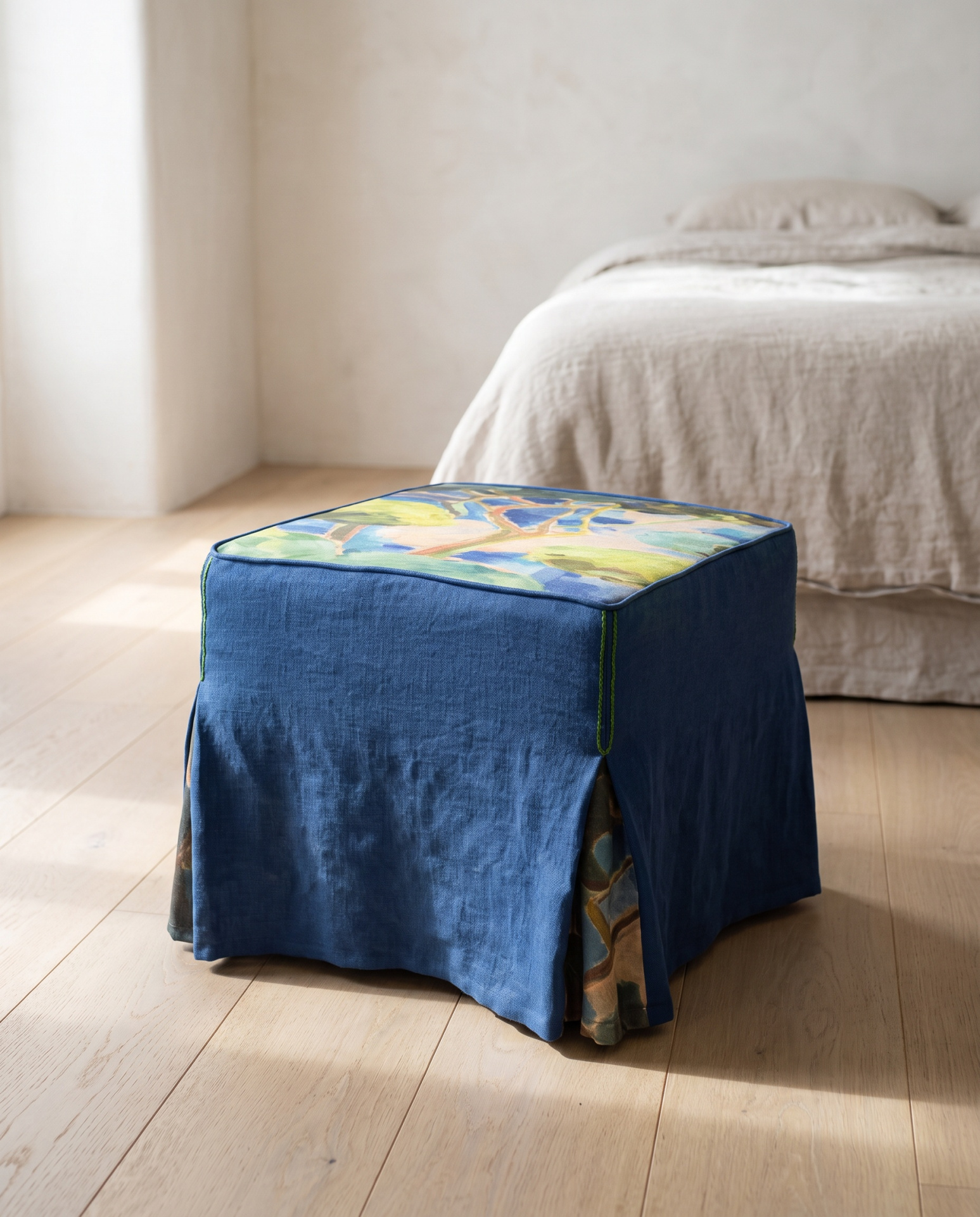 Pufe quadrado azul com padrão colorido no topo junto a uma cama com roupa bege