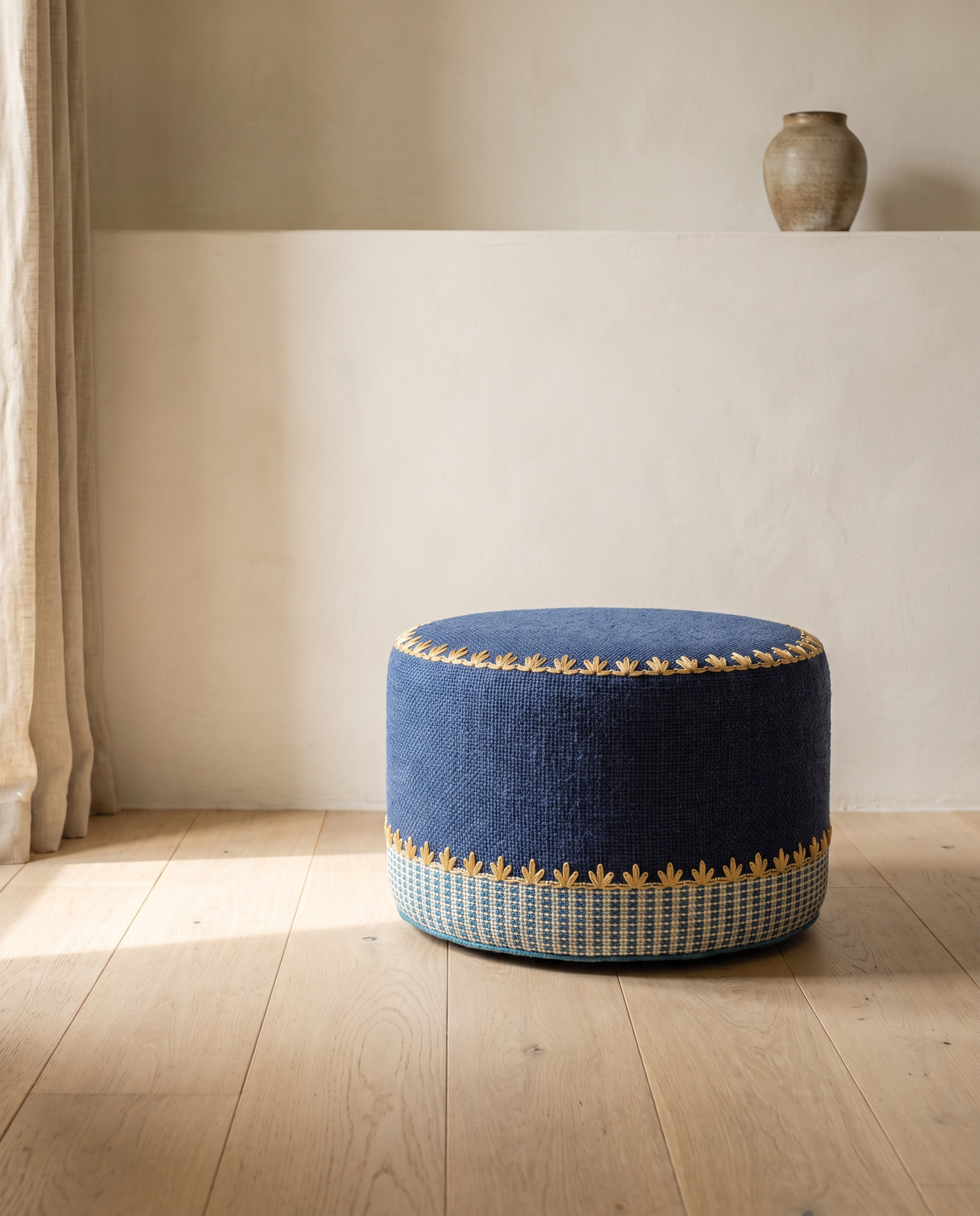 Pufe redondo azul com bordado amarelo e base quadriculada, em chão de madeira e parede clara com vaso cerâmico