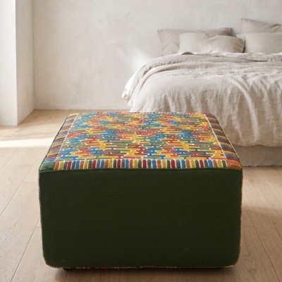 Pufe quadrado com padrão multicolorido sobre chão de madeira, cama bege ao fundo
