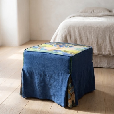 Pufe quadrado azul com padrão colorido no topo junto a uma cama com roupa bege