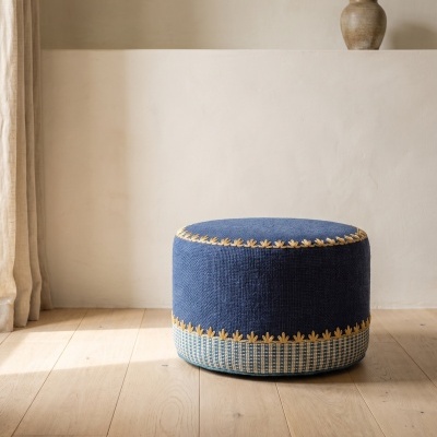 Pufe redondo azul com bordado amarelo e base quadriculada, em chão de madeira e parede clara com vaso cerâmico