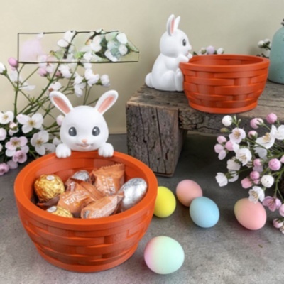 Dois cestos laranja com figuras de coelho e chocolates com ovos coloridos e flores ao redor.