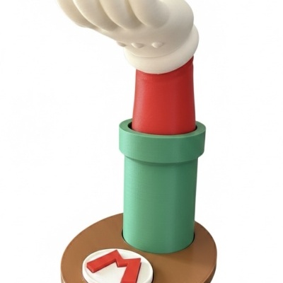 Escultura de mão branca e braço vermelho saindo de cano verde sobre base castanha com M vermelho