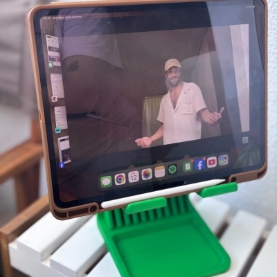 Tablet com capa castanha num suporte verde sobre mesa branca, com imagem de homem vestido de branco na tela.