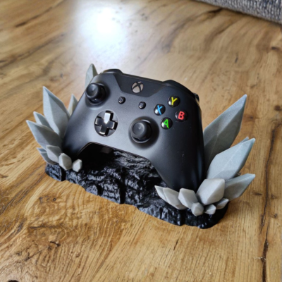Comando Xbox preto numa base decorativa cinzenta e preta em forma de cristais sobre mesa de madeira clara