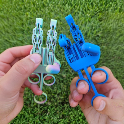 Dispositivos mecânicos plásticos azuis e verdes com design articulado, segurados por mãos, com fundo de relva verde