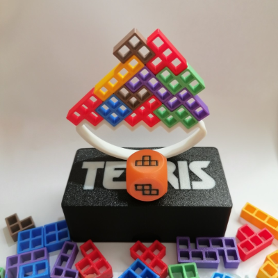 Peças de jogo de tabuleiro em plástico colorido tipo Tetris montadas em suporte branco com base preta TETRIS e dado laranja.