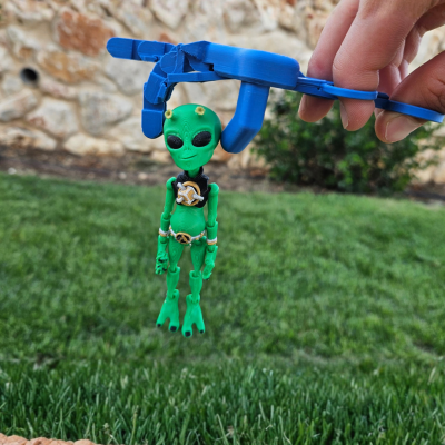 Boneco alienígena verde segurado por pinça azul com relva e muro ao fundo
