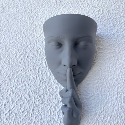 Máscara decorativa cinza de rosto com dedo indicador na boca, sobre parede branca texturada