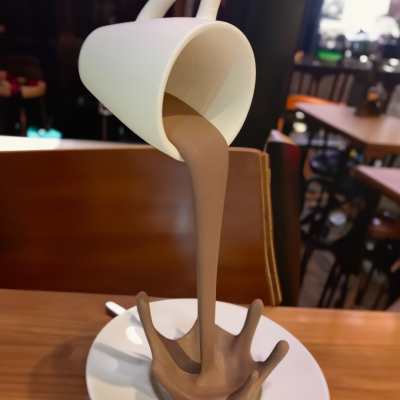 Escultura de chávena a verter líquido castanho formando splash sólido num prato branco