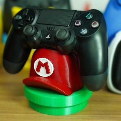 Comando de videojogo preto PlayStation num suporte em forma de chapéu vermelho com M branco e vermelho, base verde, superfície de madeira.
