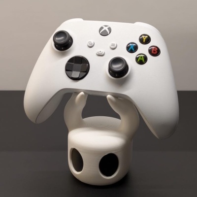 Comando Xbox branco com botões coloridos num suporte branco em forma de figura com olhos negros